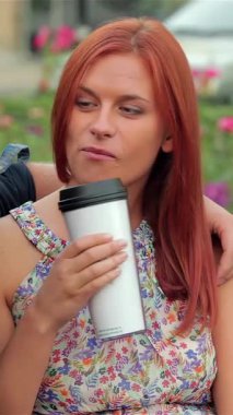 A joyful couple shares a moment of laughter on a sunlit park bench, sipping takeaway coffee while surrounded by blooming roses. Their playful interaction and relaxed smiles radiate warmth and connection, creating a delightful atmosphere in this