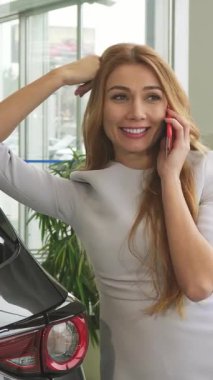 A radiant woman with long, flowing hair beams with delight as she leans against her sleek new car, fully immersed in an animated phone conversation. The bright, modern dealership showroom, adorned with luxurious vehicles, radiates an atmosphere of