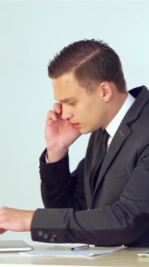 A determined young male entrepreneur, dressed in a sharp suit, intensely analyzes data on his laptop while jotting down notes and engaging in a phone conversation. This vibrant scene encapsulates the hustle of modern corporate life, highlighting his
