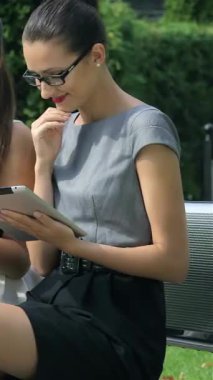 Three confident businesswomen are seated on a sleek park bench, animatedly discussing ideas while interacting with their devices. Their chic outfits and radiant smiles embody a vibrant, collaborative spirit amidst the lush greenery of an urban park