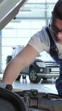 Sarışın, kendini adamış beyaz bir tamirci kaputun üzerine eğilip karmaşık motor parçalarını inceliyor. Mavi tulumlar ve şık gözlükler takmış. Parlak, modern bir garajda profesyonelliği temsil ediyor.