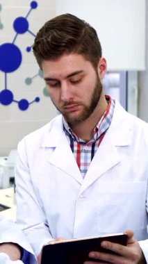 In a sleek, high-tech laboratory, two dedicated male scientists, one youthful and one seasoned, are deeply engaged in examining a tablet. The younger Caucasian scientist, clad in a crisp white lab coat, points at the screen with enthusiasm, while the