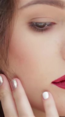 This captivating close-up features a stunning Caucasian woman gently caressing her flawless skin, embodying a moment of serene self-care. Her striking red lips and shimmering nails pop against the rich black background, radiating elegance and