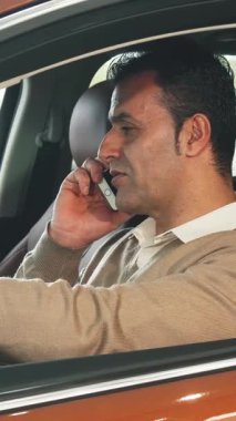 Koyu renk saçlı keyifli bir adam şık, modern bir arabada oturup telefonuyla sohbet ediyor. Bej rengi süveteri zarif kahverengi iç kısmıyla zıtlaşıyor. Gelişmişlik ve neşe saçıyor.