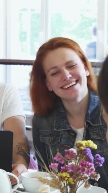 A lively scene captures a joyful young woman with vibrant red hair, sharing laughter with her handsome boyfriend and a close friend over a delightful breakfast spread. The inviting atmosphere is filled with the rich aroma of fresh pastries and coffee