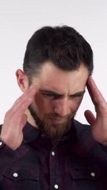A bearded young man in a dark plaid shirt stands against a soft, neutral background, rubbing his temples in a moment of intense discomfort. This striking image powerfully conveys the emotional and physical toll of a severe headache, inviting viewers