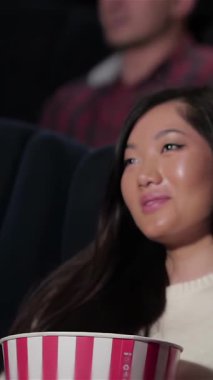 An Asian woman leans in closely to whisper sweet nothings to her girlfriend, both glowing with joy in a cozy cinema filled with laughter. Surrounded by an engaged audience, they savor buttery popcorn and refreshing soda, fully immersed in the