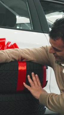 The ecstatic man beams with joy beside two sleek, brand-new car wheels, beautifully adorned with a striking red bow. With a thumbs-up and car keys in hand, he radiates excitement, capturing the thrill of a perfect automotive purchase