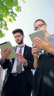 A confident businessman and a successful businesswoman joyfully celebrate exciting news while focused on their tablets in a vibrant urban park. Sunlight streams through the lush green leaves, illuminating their smiles and creating an energetic