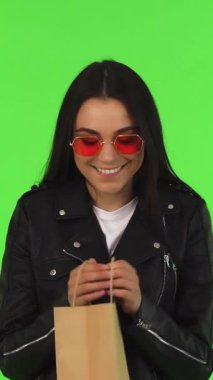 A joyful young woman with long dark hair and trendy red sunglasses radiates excitement as she eagerly peers into her shopping bag, set against a vivid green chromakey background. This dynamic scene encapsulates the thrill of retail therapy