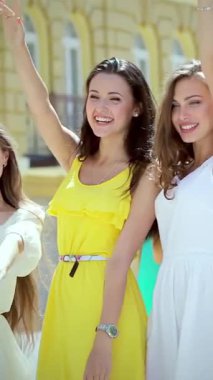 Three vibrant girls, radiating joy in colorful summer dresses, wave their shopping bags enthusiastically while strolling down a sunlit street. Their infectious laughter and bright smiles create a lively atmosphere of friendship and celebration
