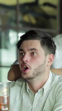 In a lively bar filled with the buzz of excitement, two young businessmen toast with frosty beer glasses, their faces radiating joy and camaraderie. One friend, animatedly shouting with enthusiasm, captures the thrill of the football match on TV