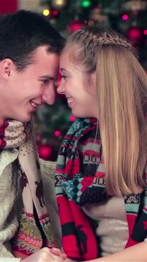A joyful couple, wrapped in snug sweaters and vibrant scarves, share tender moments while exchanging beautifully wrapped Christmas gifts in a festively adorned room. The enchanting atmosphere, illuminated by twinkling lights and a lush Christmas tree
