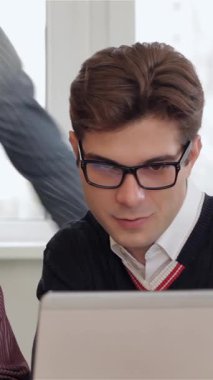 In a modern office, four young creative managers, including a stylish brunette man in glasses and a focused woman, gather around a sleek laptop, deeply engaged in animated discussion. A blond man leans in from behind, contributing to the vibrant