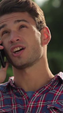 A handsome young man animatedly chats on his phone while seated on a park bench, surrounded by vibrant greenery and dappled sunlight. His joyful expression and relaxed demeanor capture the essence of outdoor leisure and meaningful connection
