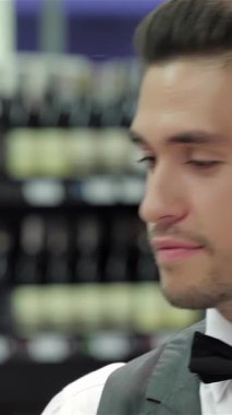 A sophisticated male sommelier in a sleek tuxedo thoughtfully examines a wine glass, his discerning gaze reflecting expertise and passion. Surrounded by an elegant backdrop of rich wine bottles on a rustic wooden shelf, the scene exudes luxury and