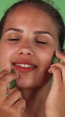 A stunning young woman joyfully massages her face with cool cucumber slices, enhancing her natural beauty against a vibrant green chromakey backdrop. This invigorating skincare ritual radiates wellness and rejuvenation, inviting viewers to indulge in