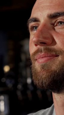 A joyful bearded man with captivating blue eyes beams with happiness as he savors a chilled beer, fully immersed in the thrilling atmosphere of a football match at a bustling pub. The rustic brick walls and warm lighting create an inviting backdrop