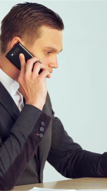 A determined young businessman, clad in a sleek suit, engages in a crucial phone call while skillfully typing on his laptop, embodying professionalism and commitment. The modern office backdrop, with its clean lines and soft lighting, amplifies the