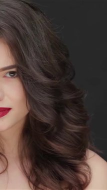 This striking close-up showcases a young Caucasian woman with flawless skin, exuding confidence and elegance against a sleek black backdrop. Her wavy, luscious hair frames her face beautifully, while bold red lips and a captivating gaze highlight her