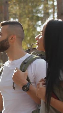 An adventurous male hiker confidently points the way through a vibrant green forest, while his girlfriend, a stunning brunette, gazes up at him with admiration. Their backpacks hint at a day filled with exploration and joy, capturing the essence of