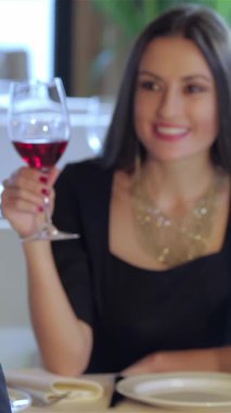 A delightful young couple shares a moment of joy, raising their glasses of rich red wine in a toast, while playfully summoning the waiter. The upscale restaurant, adorned with soft lighting and elegant roses, creates a romantic ambiance, perfect for