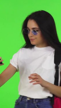 A stylish young woman with long, flowing hair and trendy sunglasses joyfully snaps selfies with her smartphone, set against a vibrant green chromakey backdrop. This dynamic scene captures the essence of modern student life, radiating her playful