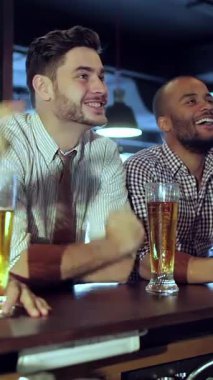 Two excited young men, dressed in smart casual attire, enthusiastically cheer while raising their frosty glasses of beer at a vibrant sports bar. The lively atmosphere buzzes with energy, laughter, and the thrill of the game, as they share joyful