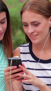Two fashionable girls, one with flowing brown hair and the other with sleek dark locks, are joyfully sharing music on a smartphone while sitting on a rustic wooden bench in a lively city park. Their relaxed smiles and trendy summer outfits radiate a