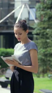 A confident businesswoman radiates positivity in a sleek black suit, smiling brightly at the camera. Behind her, colleagues animatedly discuss ideas while holding tablets, set against a lush green backdrop of a modern urban landscape, embodying