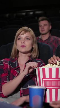 A vibrant group of young friends is fully engaged in a captivating film at a cozy cinema, their laughter echoing in the air. With oversized popcorn buckets and colorful drinks in hand, the joyful atmosphere is alive with excitement as they bond over