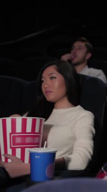 A vibrant group of young friends is fully engaged in a captivating film at the cinema, their faces lit with joy and anticipation. With oversized popcorn buckets and refreshing drinks, the cozy atmosphere buzzes with laughter and excitement, creating