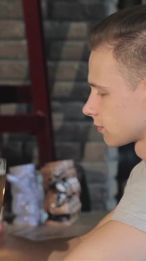 A dapper young Caucasian man enjoys a refreshing golden beer at a chic bar, his relaxed demeanor radiating contentment. The inviting atmosphere, accentuated by a rustic brick wall and soft lighting, sets the perfect scene for unwinding and savoring