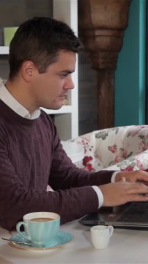 A dapper Caucasian man beams with joy as he types on his laptop, savoring a rich espresso in a cozy cafe adorned with floral decor. The bright, inviting space, filled with natural light and stylish furnishings, creates an ideal backdrop for