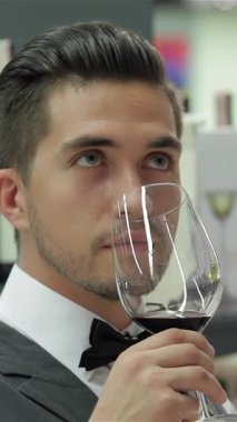 Zarif genç bir erkek şarap garsonu, şık bir yelek ve papyon giyer, zarif bir şekilde bir bardak koyu kırmızı şarap tutar, zengin, kompleks aromalarını içine çeker, odaklanmış bir minnettarlık bakışı ile. Bir dizi sofistike şarap şişesiyle çevrili bu sahne...