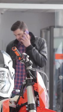 Şık, esmer bir adam şık turuncu bir motosikletin yanında duruyor, canlı bir galeride derin bir telefon görüşmesi yapıyor. Parmakları motosiklet koltuğunu hafifçe sıyırdı. Heyecan dolu dünyadaki tutkusunu ve uzmanlığını yansıttı.