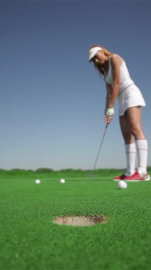 Şık beyaz elbiseli, çarpıcı kızıl saçlı bir kadın, üç golf topuna ustalıkla, gür, canlı yeşil bir sahada vururken, şiddetle konsantre oluyor. Sahne kararlılık ve kesinlik saçıyor, toplar deliğin kenarında sallanıyor.