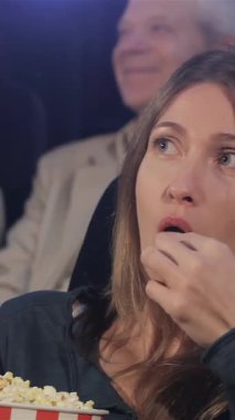 Orta yaşlı bir kadın, gözleri dört açıkta, çeşitli izleyicilerle dolu canlı bir sinemada bir kova patlamış mısırdan hoşlanıyor. Atmosfer heyecan ve kahkahalarla dolu. Kendini sinematik deneyime kaptırıyor.