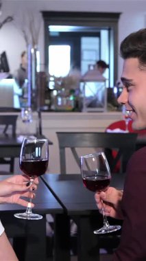 Hoş bir yakın plan, genç bir çifti şık bir restoranda kadehlerini kırmızı şarapla tokuştururken, gülümsemeleri sıcaklık ve bağlantı yayarken yakalıyor. Sırt sırta oturan beyaz erkek ve kadın, samimi bir atmosfer yaratıyor.