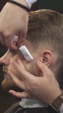 Bu yakın plan çekimde yakışıklı, bakımlı sakallı genç bir adam, profesyonel bir berber gibi yüzünü ustura ile titizlikle tıraş ediyor. Klasik berber dükkânı havası, sanatsal ve sanatsal deneyimleri ön plana çıkarıyor.