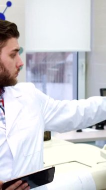 Zarif, yüksek teknolojili bir laboratuvarda, şık sakallı odaklanmış beyaz bir adam ekranı işaret ederken titizlikle tabletteki verileri analiz ediyor. Sahne, düşünceli, gri saçlı, üst düzey bir laboratuvar çalışanı tarafından zenginleştirildi.