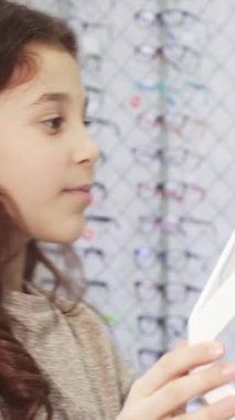 Dalgalı kahverengi saçlı hoş küçük bir kız gözlükçüde hareketli bir güneş gözlüğü seçimi yaparken neşe saçıyor. Göz kamaştırıcı renkli çerçeveler ve eğlenceli tasarımlar hayal gücünü ateşliyor ve onu alışveriş yapmaya zorluyor.
