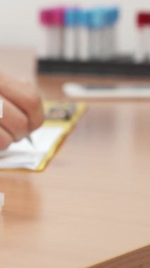 Renkli numunelerle dolu canlı bir kan test tüpü koleksiyonu, sade ahşap bir masanın üzerinde duruyor, sağlık hizmetlerindeki teşhisin kritik rolünü sembolize ediyor. Arka planda, koyu saçlı ve sakallı kendini adamış beyaz bir erkek doktor var.