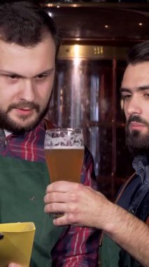 İki kendini adamış erkek bira üreticisi, biri sakallı, parlak paslanmaz çelik ekipmanlarla çevrili, canlı altın içkilerini dikkatle analiz ediyor. Odaklanmış ifadeleri ve canlı tartışmaları sanatsal ve takım çalışmasının içeriğini vurguluyor