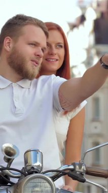 Neşeli genç bir çift, hayat dolu şehir sokaklarında scooter sürerken, erkek heyecan verici bir manzarayı işaret ederken kadın onu sıkıca kucaklıyor. Bu dinamik şehir manzarası macera ve sevgi saçıyor.