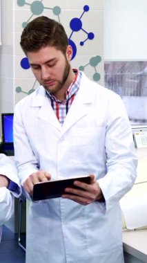 Kendini adamış esmer bir stajyer gri saçlı üst düzey meslektaşı ile lüks, modern bir laboratuvarda tabletten görüşlerini paylaşıyor. Canlı sahne, gelişmiş laboratuvar ekipmanları ve renkli malzemelerle çevrelenmiş işbirliklerinin altını çiziyor.