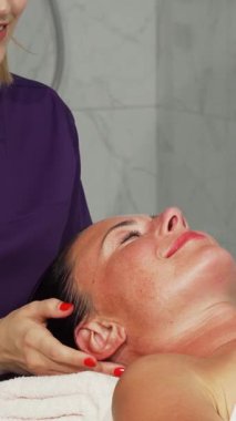 Huzurlu bir spa ortamında, yetenekli bir terapist mutlu ve rahat bir kadının kafasına nazikçe masaj yapar, gözleri kapalı gülümseyen, saf neşe saçan bir kadın. Yumuşak ışıklandırma ve sakinleştirici renkler sahneyi kaplıyor. Huzur vahası yaratıyor.