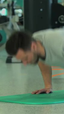 Ortadoğu kökenli kendini adamış genç bir adam güçlü ve kararlı bir şekilde parlak yeşil bir egzersiz minderinin üzerinde şınav çekiyor. Çeşitli çalışma ekipmanlarıyla dolu enerjik spor salonu arka planı,