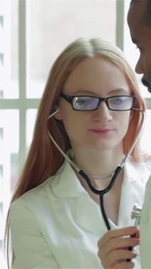 Güneşli bir klinikte, kendini işine adamış bir kadın doktor steteskopuyla genç bir adamın göğsünü dikkatle dinler ve titiz bir muayeneden geçer. Sıcak gülüşü ve profesyonel tavırları güven verici bir atmosferi teşvik ediyor.