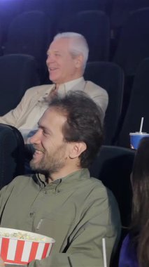 Canlı bir seyirci bir komedi filmine dalmış, kahkahaları loş sinemada yankılanıyor. Elinde bir kase patlamış mısırla genç bir adam, bir yoldaşla hareketli tepkileri paylaşıyor, neşe ve bağlantı dolu bir atmosfer yaratıyor.